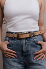Woman wearing a white ribbed tank top, blue denim jeans, and a tan leather belt with a gold buckle.